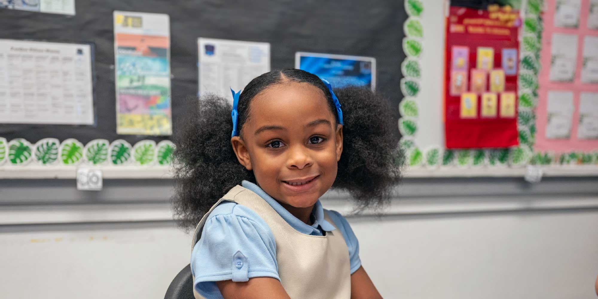 Smiling young student with pigtails and blue bows in a classroom.